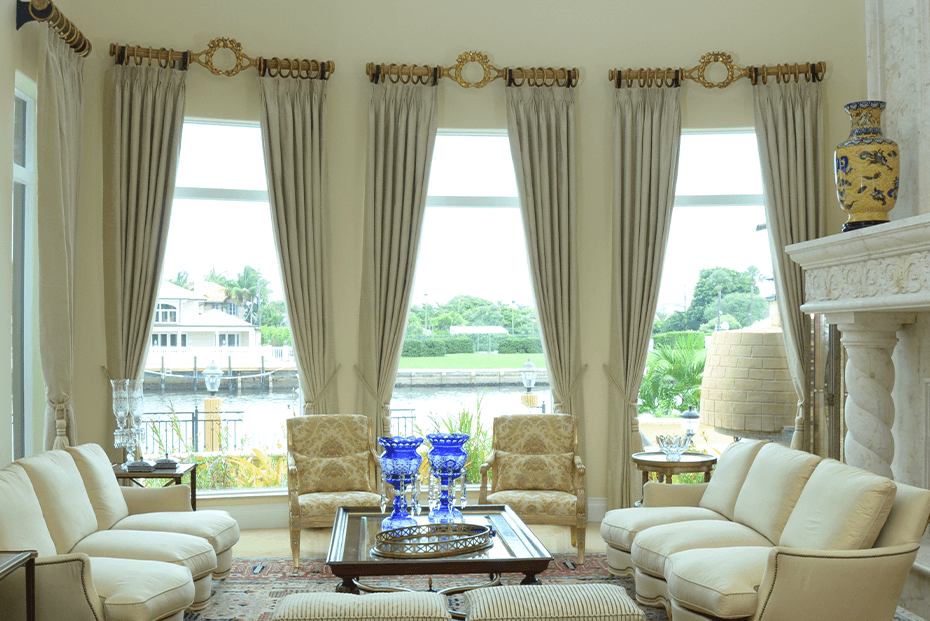 Formal living room with beige drapery and ornate gold curtain rods, framing large windows.